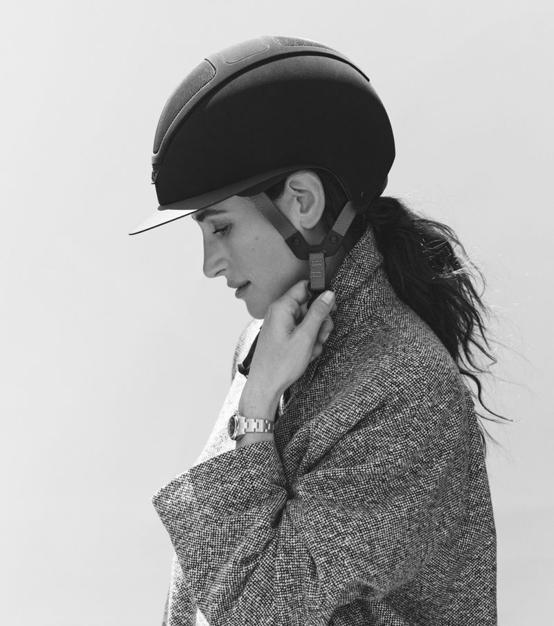 Profile portrait of a woman wearing an equestrian helmet and textured tweed coat, adjusting her chin strap against a plain background