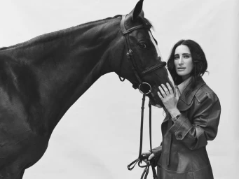 Woman in a leather trench coat standing next to a dark horse against a plain white background