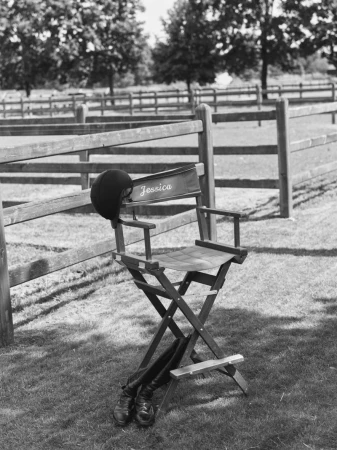 Black and white photograph of an equestrian director chair with a riding helmet and boots in a paddock.