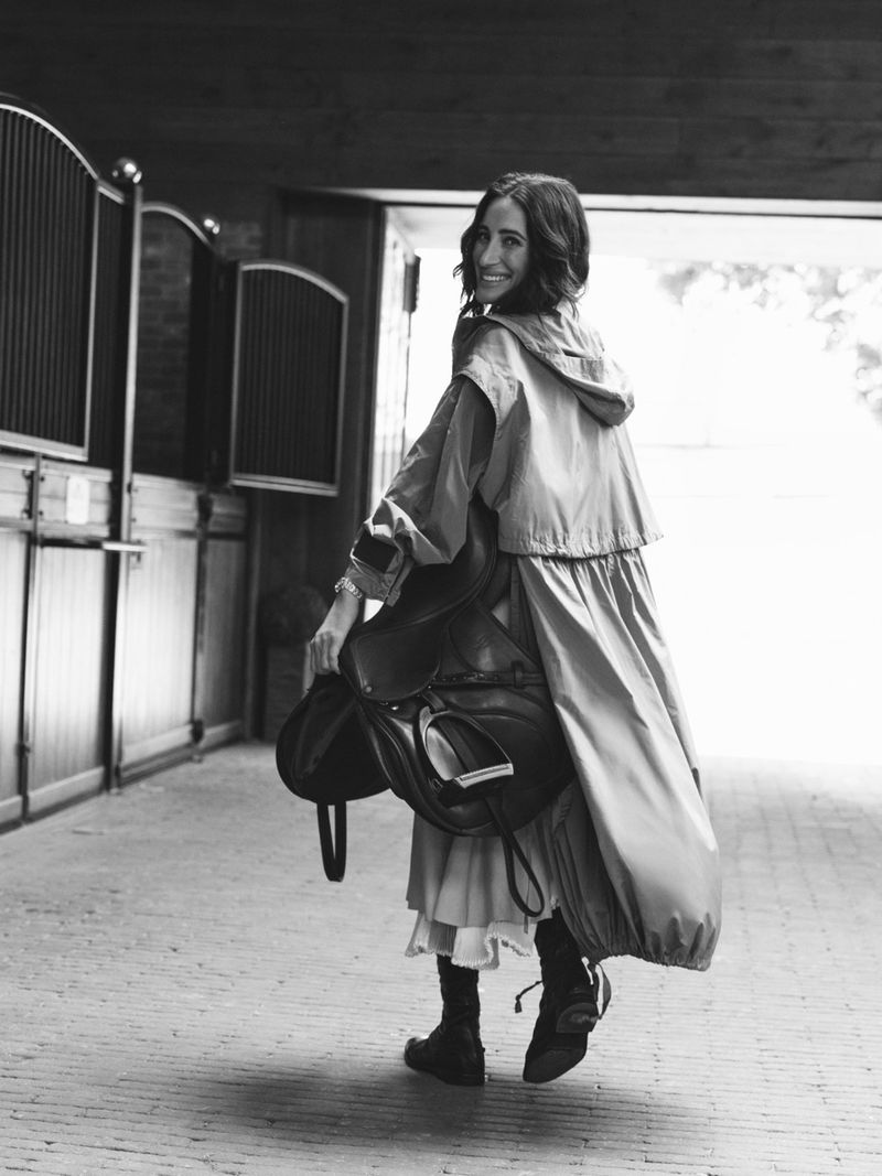 Woman in a long hooded coat carrying a leather horse saddle through a stable aisle, looking back smiling.