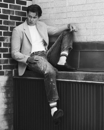 Bill Skarsgård wearing a blazer, white t-shirt, and jeans, seated outdoors against a brick wall for Fantastic Man.