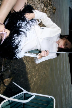 Model seated outdoors near water wearing white jacket and feather skirt under black umbrella