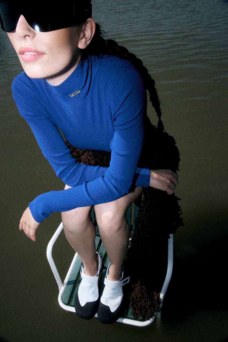 Model in oversized blue turtleneck and sunglasses sitting on a white framed chair above water.