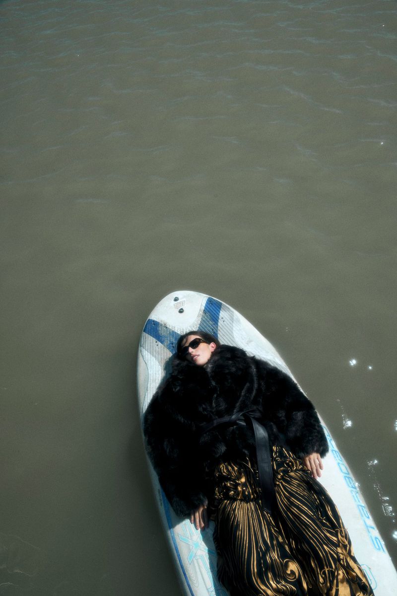 Model lying on a paddleboard in murky water wearing a black fur jacket and patterned gold skirt