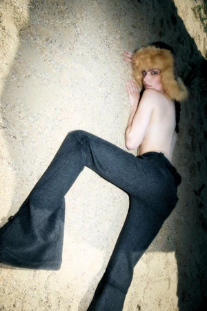 Model wearing dark trousers and a furry hat lying on sandy terrain in direct harsh lighting.