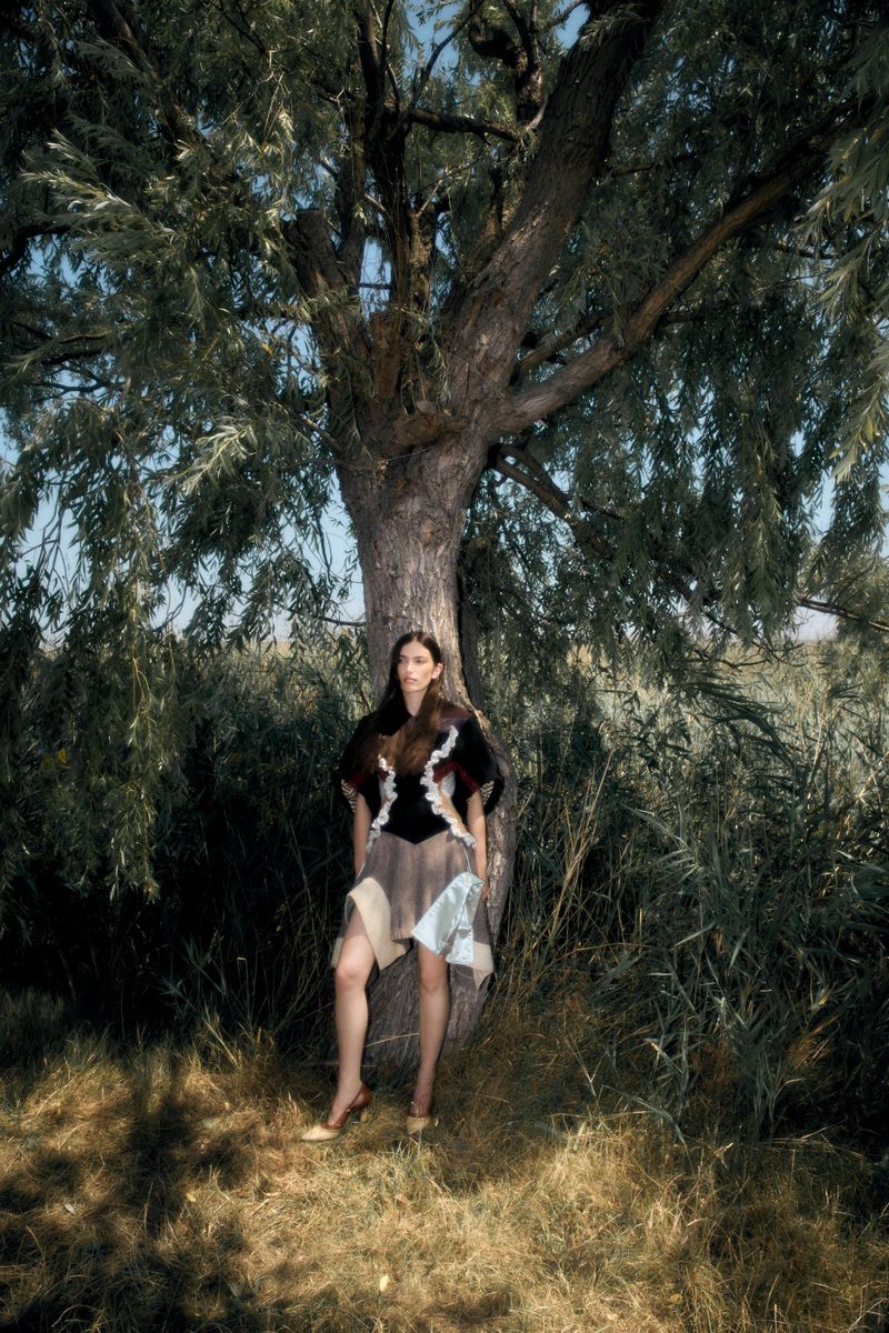Model leaning against a large tree in a wooded landscape wearing a panelled skirt and dark top