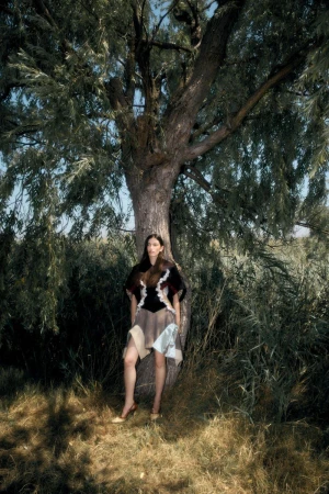 Model leaning against a large tree in a wooded landscape wearing a panelled skirt and dark top