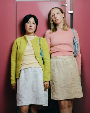 Two women standing side by side against a pink partition, wearing colourful knitwear and skirts for W Youth Pen15 Forever project.