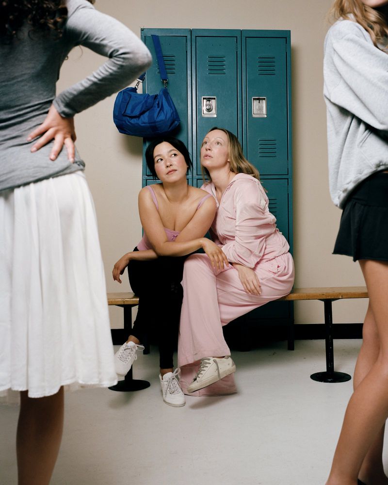 Maya Erskine and another woman seated together on a wooden bench in front of teal school lockers with standing figures framing the foreground.