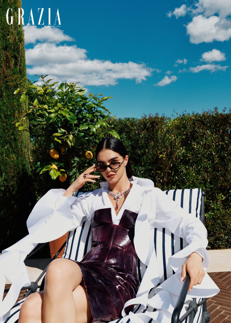 Model wearing dark sunglasses and a structured strapless dress seated on an outdoor lounger beneath a bright blue sky.