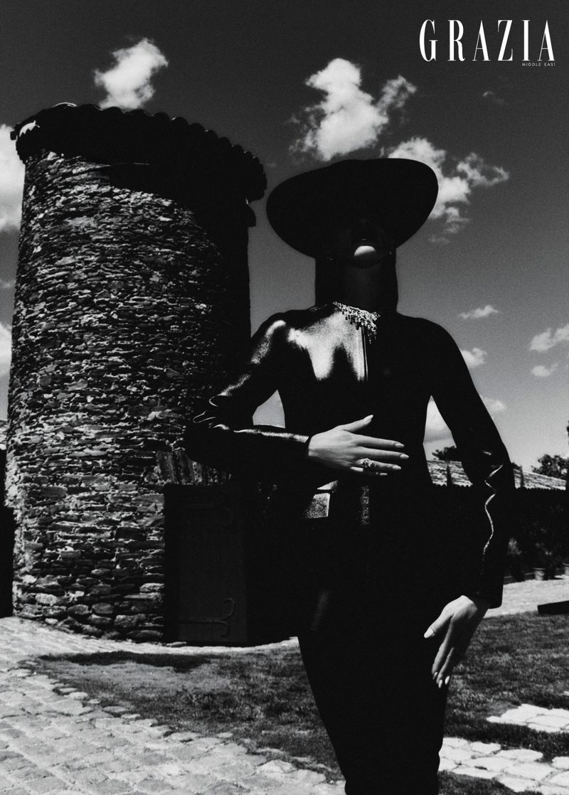 Model in dark glossy bodysuit and wide hat standing near stone tower under cloudy sky for Grazia Middle East