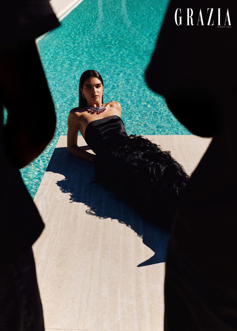 Woman in a black strapless gown reclining by a turquoise swimming pool framed by dark shadows.