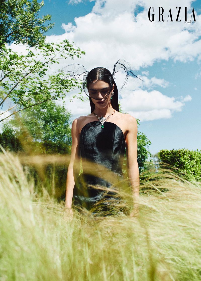 Model wearing a black dress and winged headpiece standing in a grassy field under a cloudy sky for Grazia Middle East.