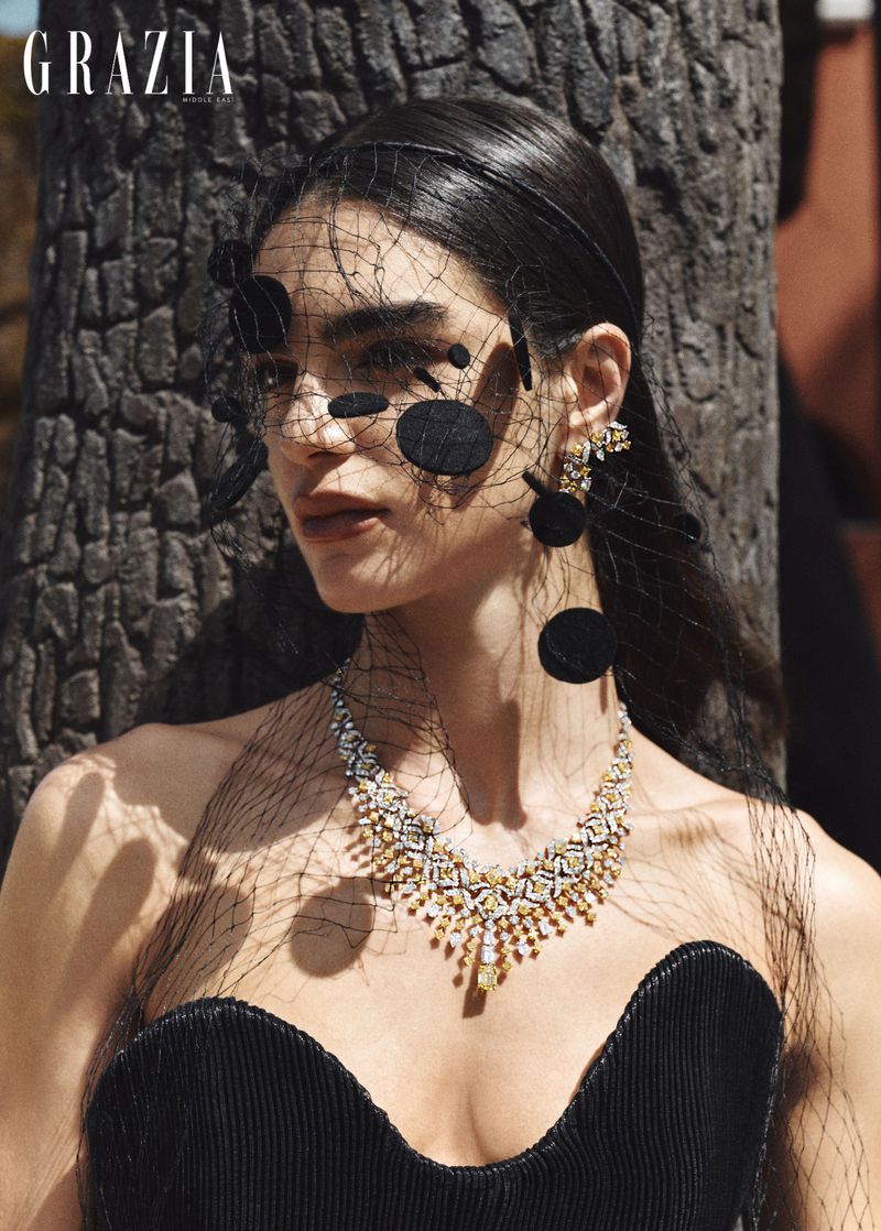 Model wearing a black dress, elaborate diamond necklace, and a black net veil with circular appliqués against a textured tree trunk.