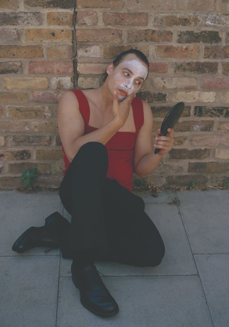 Frank Dillane seated on pavement applying white face makeup while looking into a handheld mirror before a brick wall