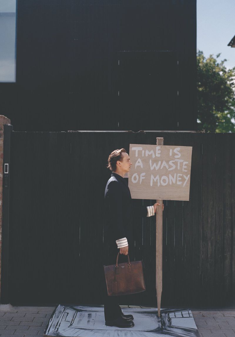 Frank Dillane standing in profile holding a sign that reads time is a waste of money and a brown briefcase