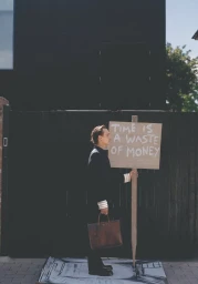 Frank Dillane standing in profile holding a sign that reads time is a waste of money and a brown briefcase