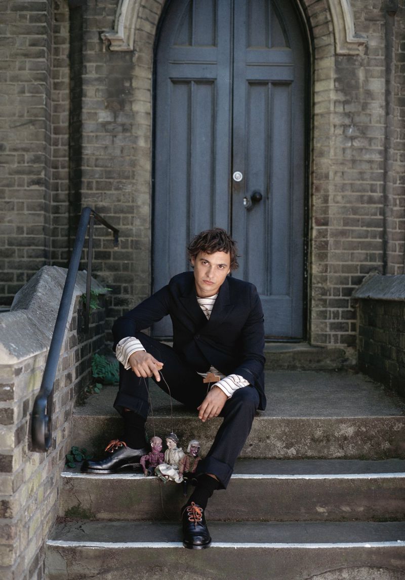 Frank Dillane sitting on stone steps in a dark suit holding marionette puppets for Crash Magazine editorial.