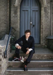 Frank Dillane sitting on stone steps in a dark suit holding marionette puppets for Crash Magazine editorial.