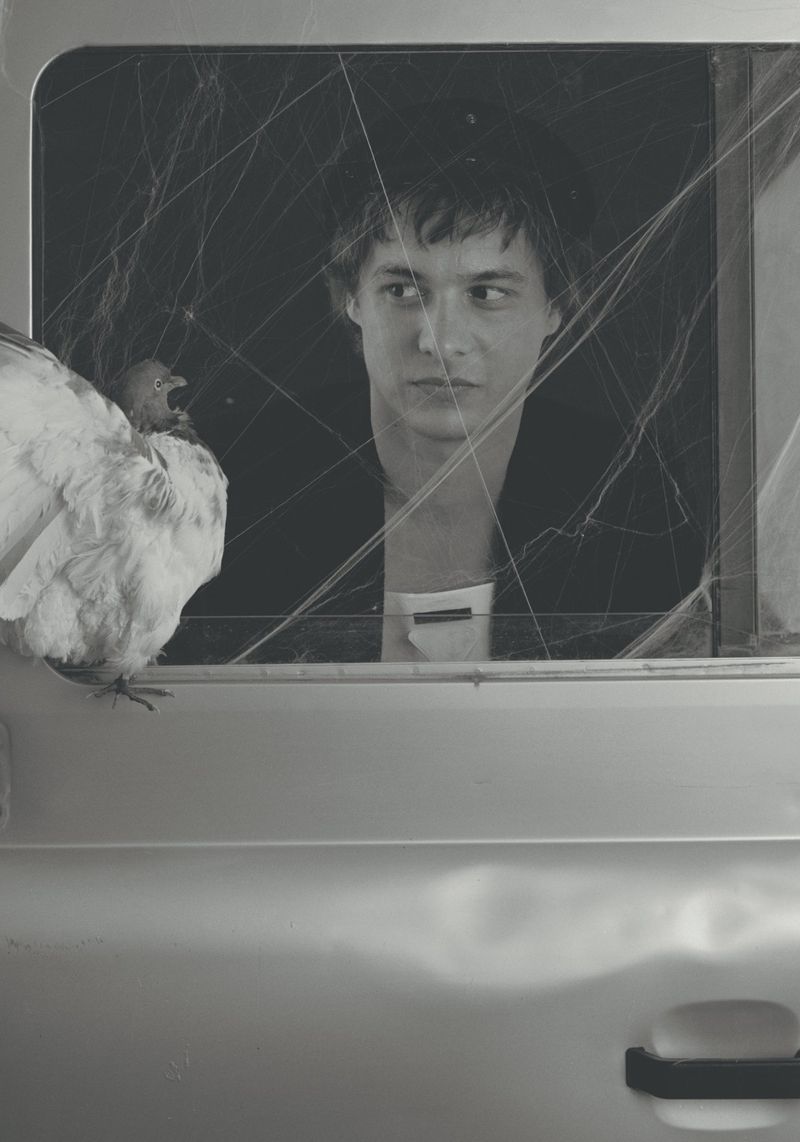 Frank Dillane looking through a car window covered in spiderwebs with a pigeon perched outside in a black and white portrait.