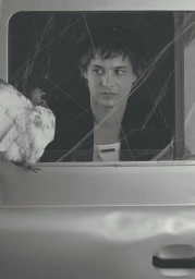 Frank Dillane looking through a car window covered in spiderwebs with a pigeon perched outside in a black and white portrait.