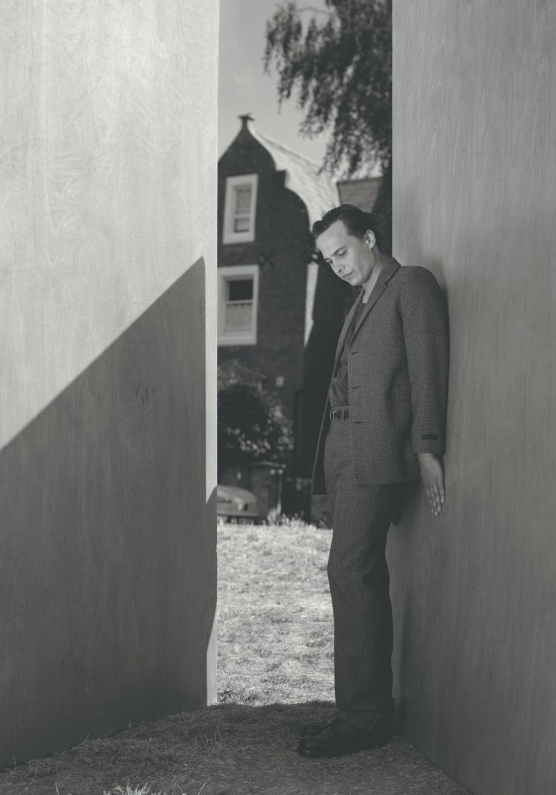 Frank Dillane wearing a tailored suit leaning against a wooden wall for Crash Magazine