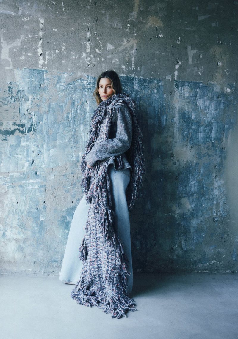 Jessica Biel standing against a textured concrete wall wearing a textured fringe wrap and wide-leg trousers.