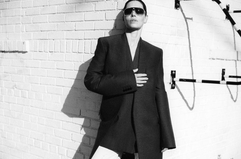Jessica Biel wearing dark sunglasses and an oversized dark blazer leaning against a white brick wall outdoors.
