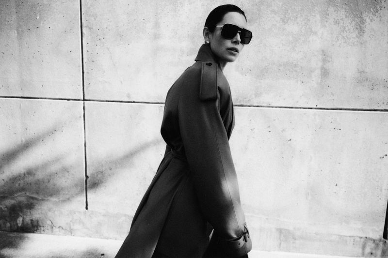 Jessica Biel wearing oversized sunglasses and a long belted coat against a concrete wall in a black and white portrait.