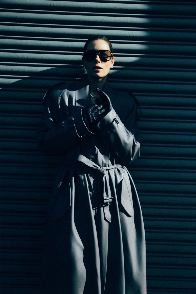 Jessica Biel wearing oversized sunglasses and a long belted coat posing against a corrugated metal shutter.