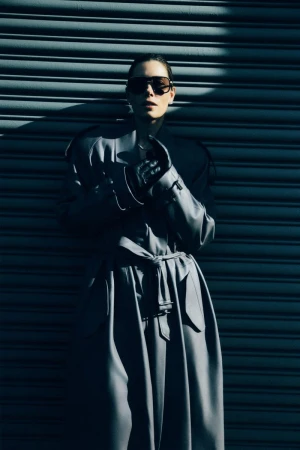Jessica Biel wearing oversized sunglasses and a long belted coat posing against a corrugated metal shutter.