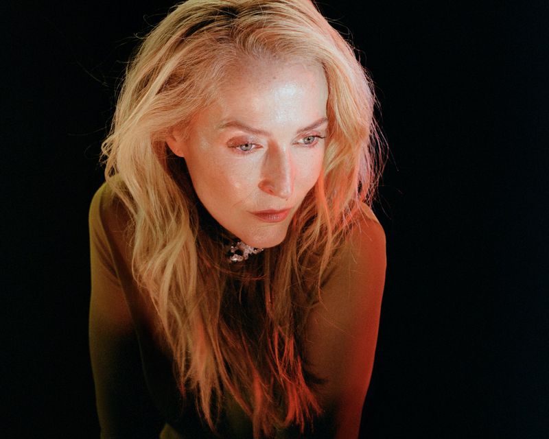 Portrait of actor Gillian Anderson with long blonde hair against a dark background with warm side lighting for Paper Magazine.