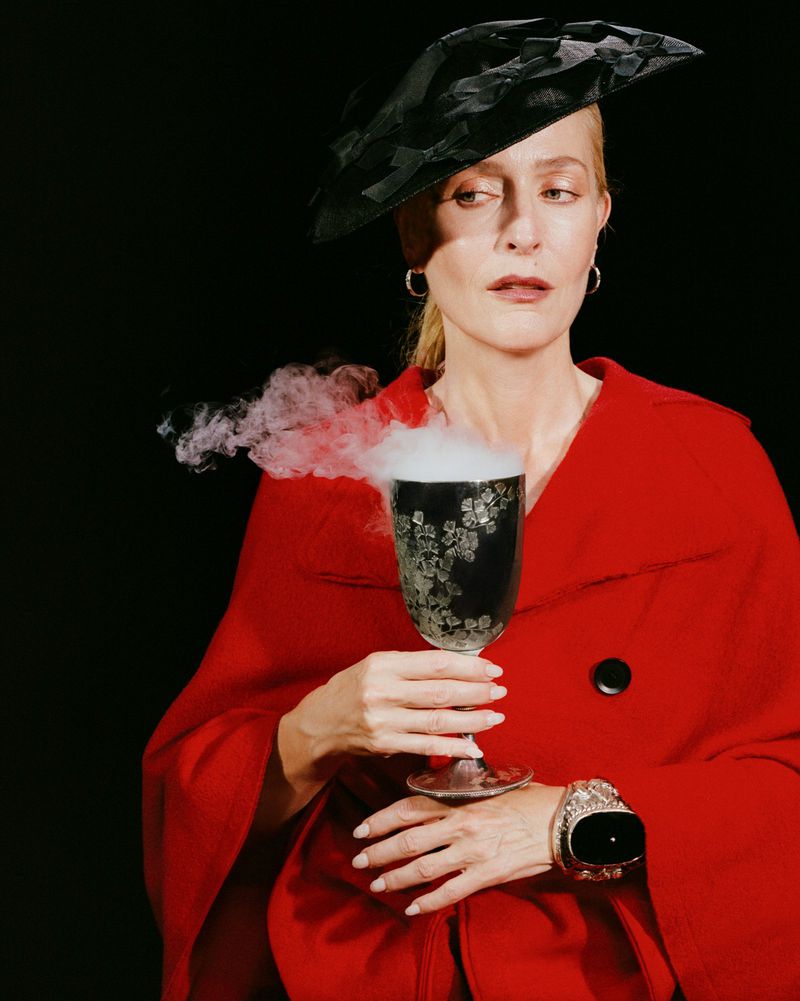 Gillian Anderson wearing a red coat and black hat holding a smoking chalice against a dark background for Paper Magazine