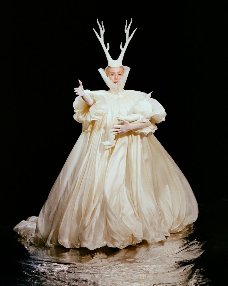 Gillian Anderson wearing a white gown and antler headpiece holding a baby doll against a black background