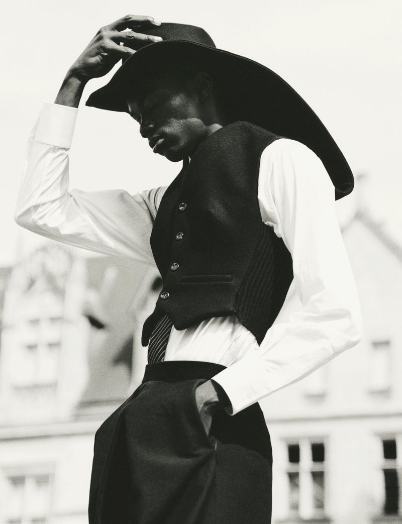 Black model adjusting a wide-brimmed hat wearing a white button-down shirt and dark vest against a building exterior.