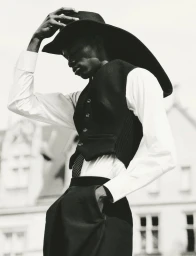 Black model adjusting a wide-brimmed hat wearing a white button-down shirt and dark vest against a building exterior.