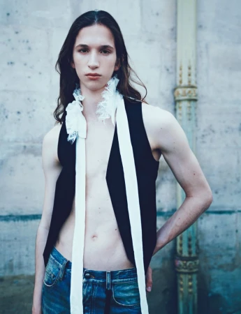 Model with long dark hair wearing a black vest and denim jeans standing outdoors against a stone wall