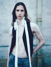 Model with long dark hair wearing a black vest and denim jeans standing outdoors against a stone wall