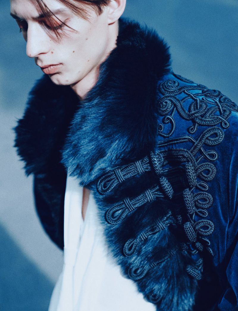 Close-up portrait of a male model wearing a blue velvet coat with faux fur collar and braided frog closures