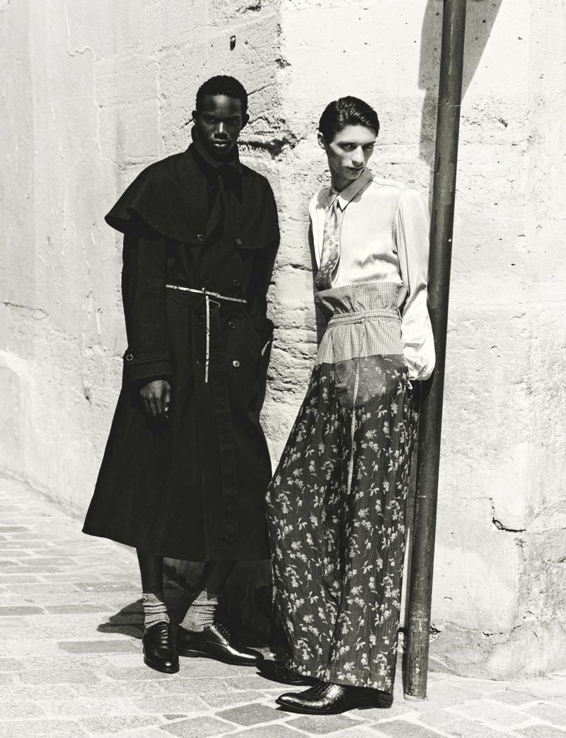 Two male models posing outdoors against a textured stone wall in black and white fashion editorial photography