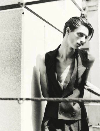 Black and white portrait of a male model in a dark vest posing outdoors near architectural walls.