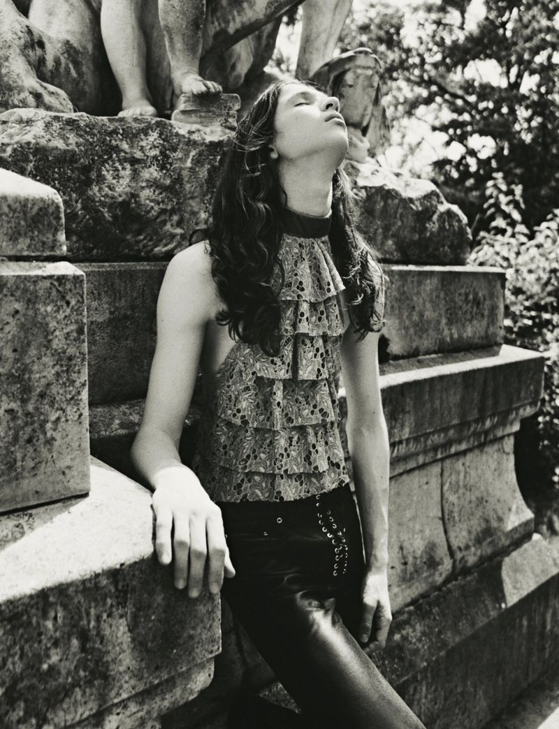 Model with long dark hair wearing a ruffled top and dark trousers leaning against large stone architectural structure in black and white.