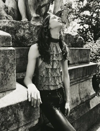 Model with long dark hair wearing a ruffled top and dark trousers leaning against large stone architectural structure in black and white.