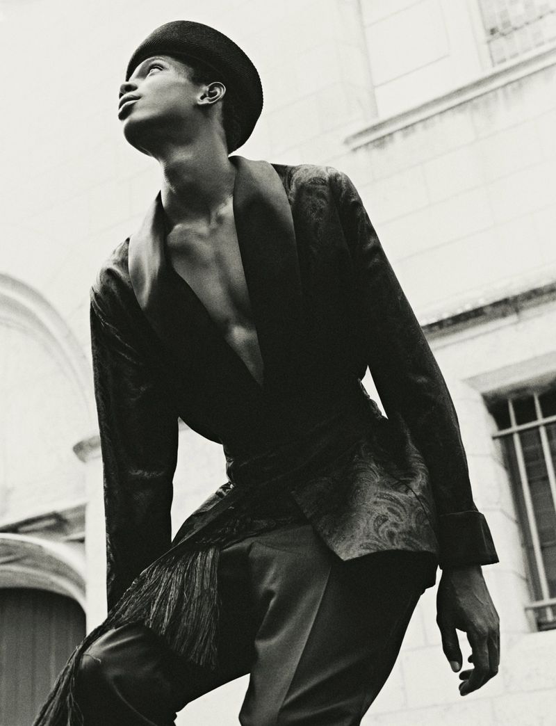 Model in dark patterned jacket and cap looking upward against an architectural stone building exterior