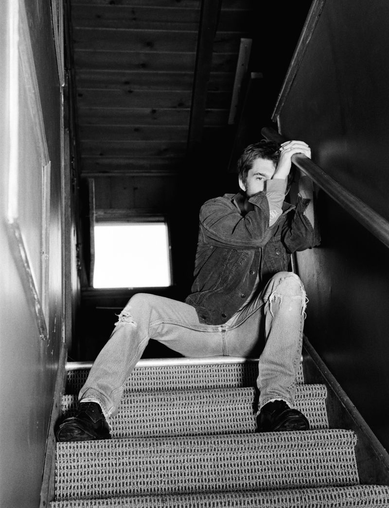 Young man in dark jacket and distressed jeans sitting on indoor carpeted stairs holding hand to face