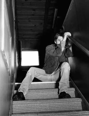 Young man in dark jacket and distressed jeans sitting on indoor carpeted stairs holding hand to face