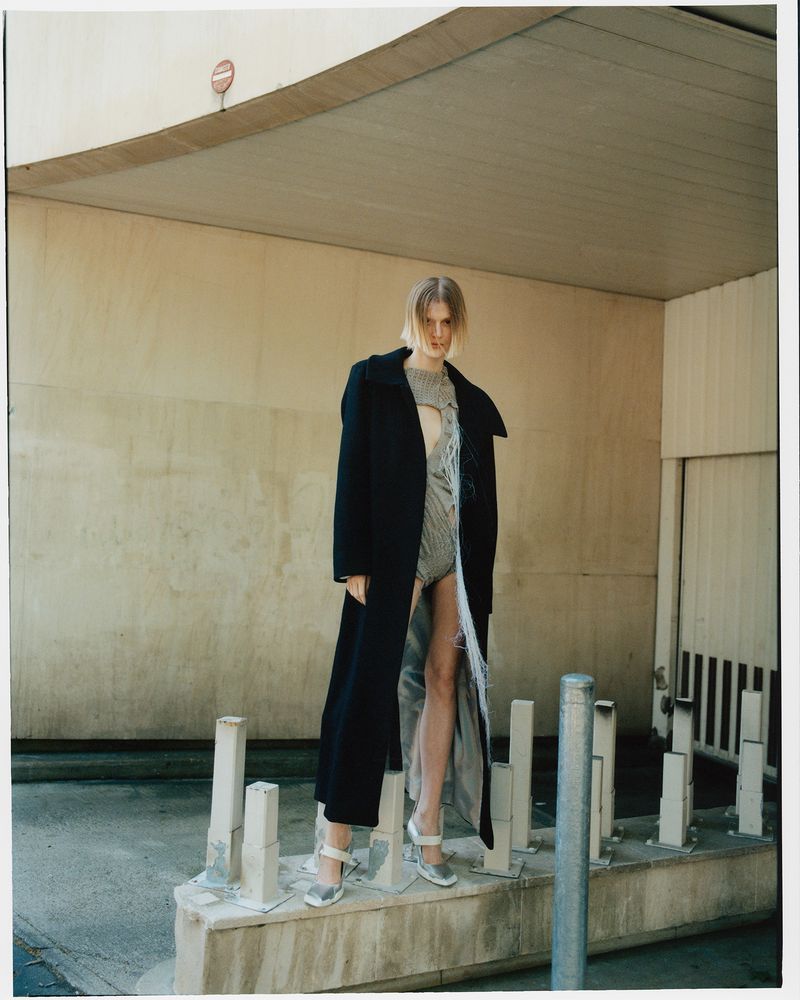 A fashion model in a gray patterned bodysuit and long black coat poses in an urban setting, highlighted by neutral colors and geometric architecture.