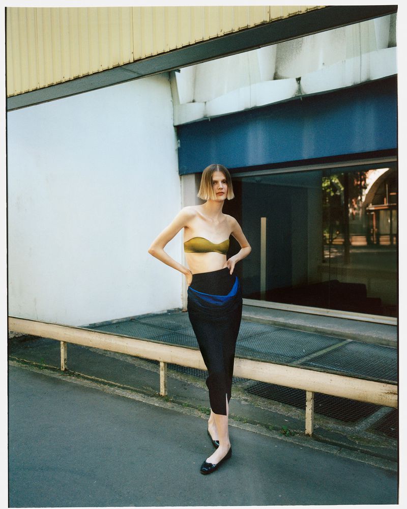A slender adult female model poses outdoors in a strapless gold top and black skirt, standing in front of a commercial building on an overcast day.
