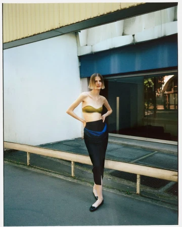 A slender adult female model poses outdoors in a strapless gold top and black skirt, standing in front of a commercial building on an overcast day.