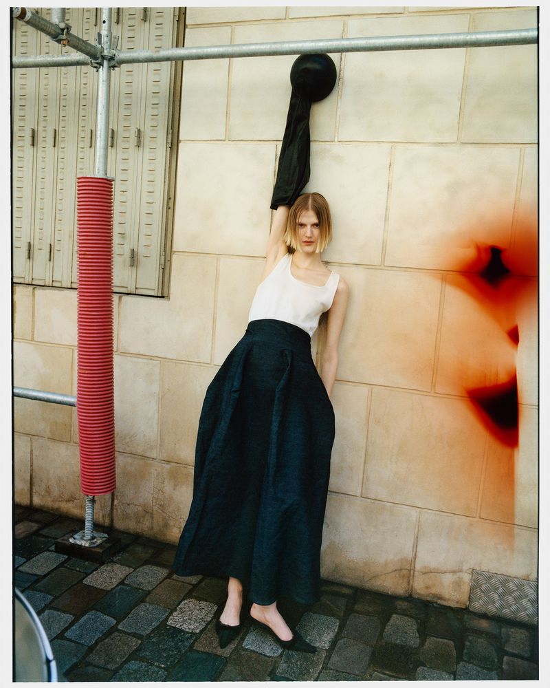 A fashionable woman in a white top and dark skirt poses against a stone wall outdoors with scaffolding and muted orange details.